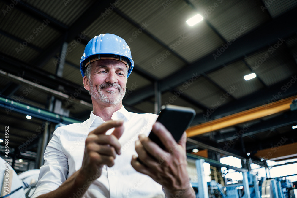 Smiling male engineer with hardhat using mobile phone at metal industry Smiling male engineer with hardhat using mobile phone at metal industry