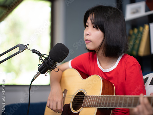 Happy cheerful pretty smiling of portrait a Asian girl vocalist with a Guitar recording a song front of microphone in living room.