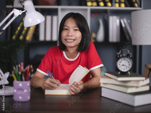 Happy smiling school girl pupil studying at home sitting at desk. Smart cute kid primary school student writing in exercise book doing homework, learning at table.