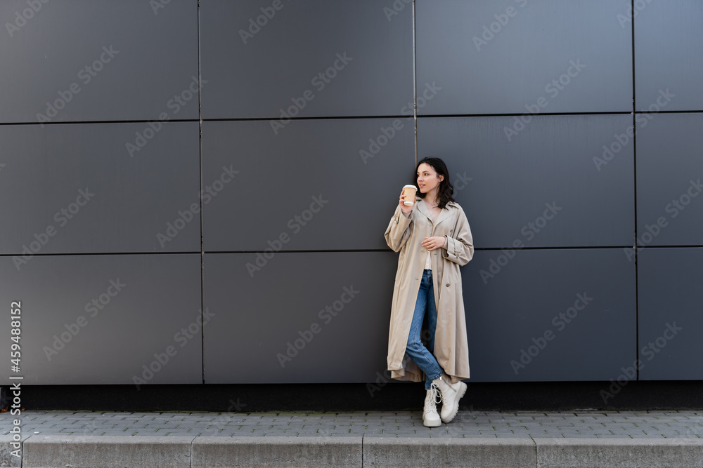 young woman in long raincoat standing with coffee to go near grey wall