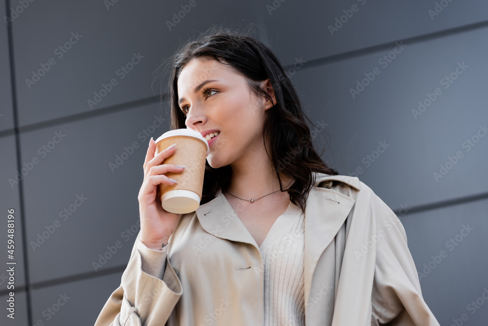 young stylish woman drinking coffee to go while looking away near grey wall young stylish woman drinking coffee to go while looking away near grey wall