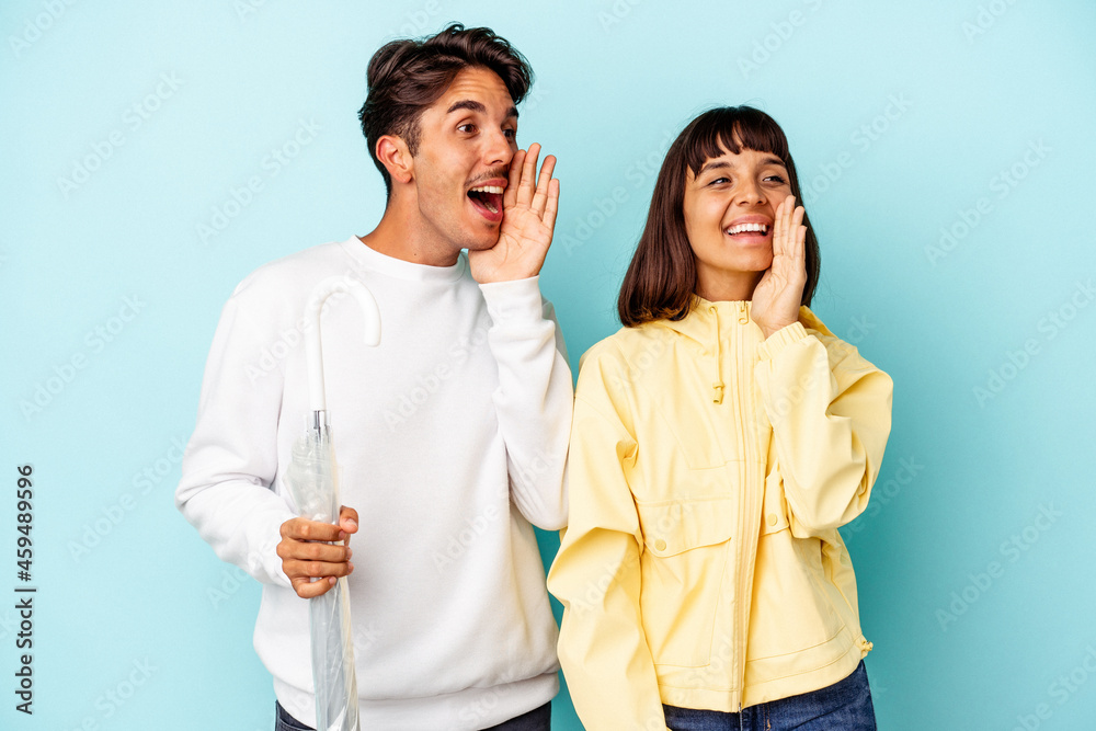 Young mixed race couple holding umbrella isolated on blue background shouting and holding palm near opened mouth. Young mixed race couple holding umbrella isolated on blue background shouting and holding palm near opened mouth.