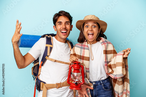 Young mixed race hiker couple isolated on blue background receiving a pleasant surprise, excited and raising hands.