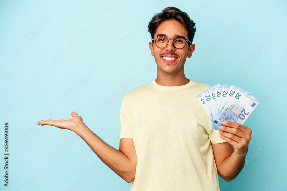Young mixed race man holding bills isolated on blue background showing a copy space on a palm and holding another hand on waist. Young mixed race man holding bills isolated on blue background showing a copy space on a palm and holding another hand on waist.