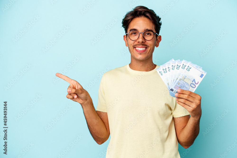 Young mixed race man holding bills isolated on blue background smiling and pointing aside, showing something at blank space. Young mixed race man holding bills isolated on blue background smiling and pointing aside, showing something at blank space.