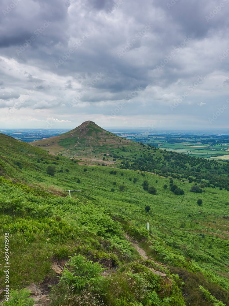 Roseberry Topping under a clouded sky, looking from the east side path with a patchwork of Teesside fields behind to the horizon.