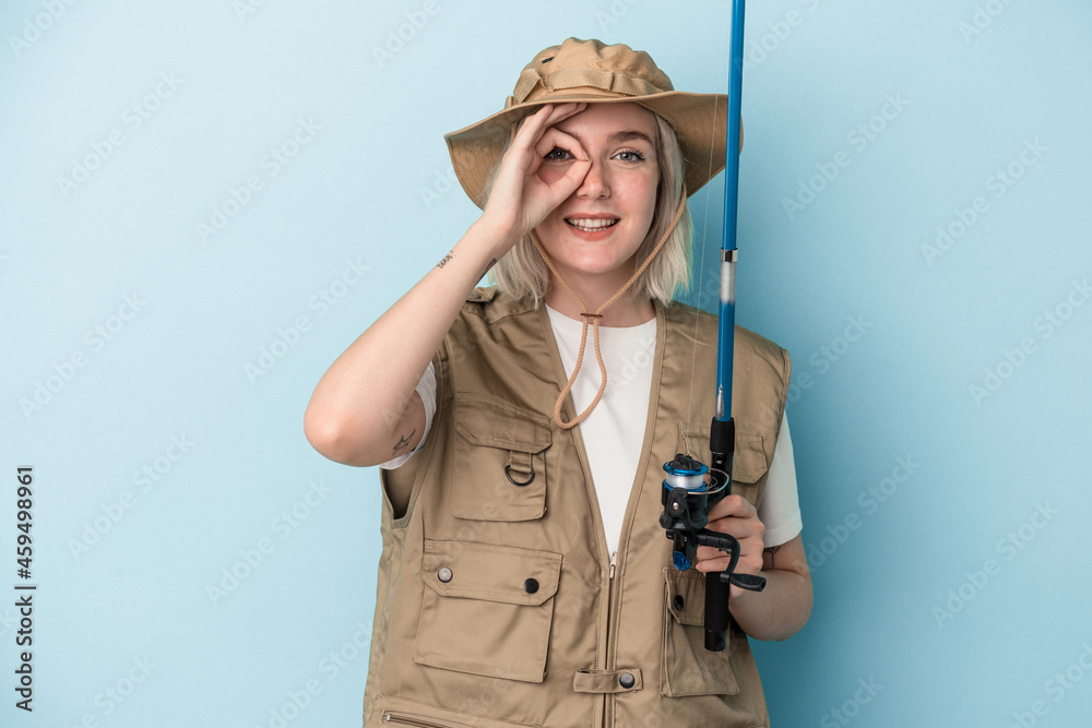 Young caucasian fisherwoman holding a rod isolated on blue background excited keeping ok gesture on eye. Young caucasian fisherwoman holding a rod isolated on blue background excited keeping ok gesture on eye.