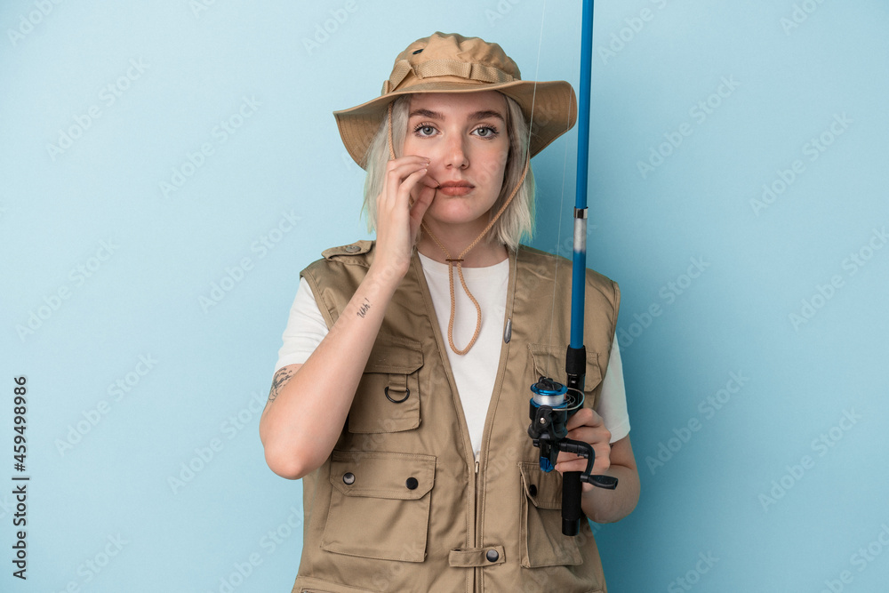 Young caucasian fisherwoman holding a rod isolated on blue background with fingers on lips keeping a secret. Young caucasian fisherwoman holding a rod isolated on blue background with fingers on lips keeping a secret.