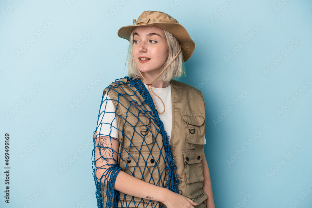 Young caucasian fisherwoman holding a net isolated on blue background looks aside smiling, cheerful and pleasant. Young caucasian fisherwoman holding a net isolated on blue background looks aside smiling, cheerful and pleasant.