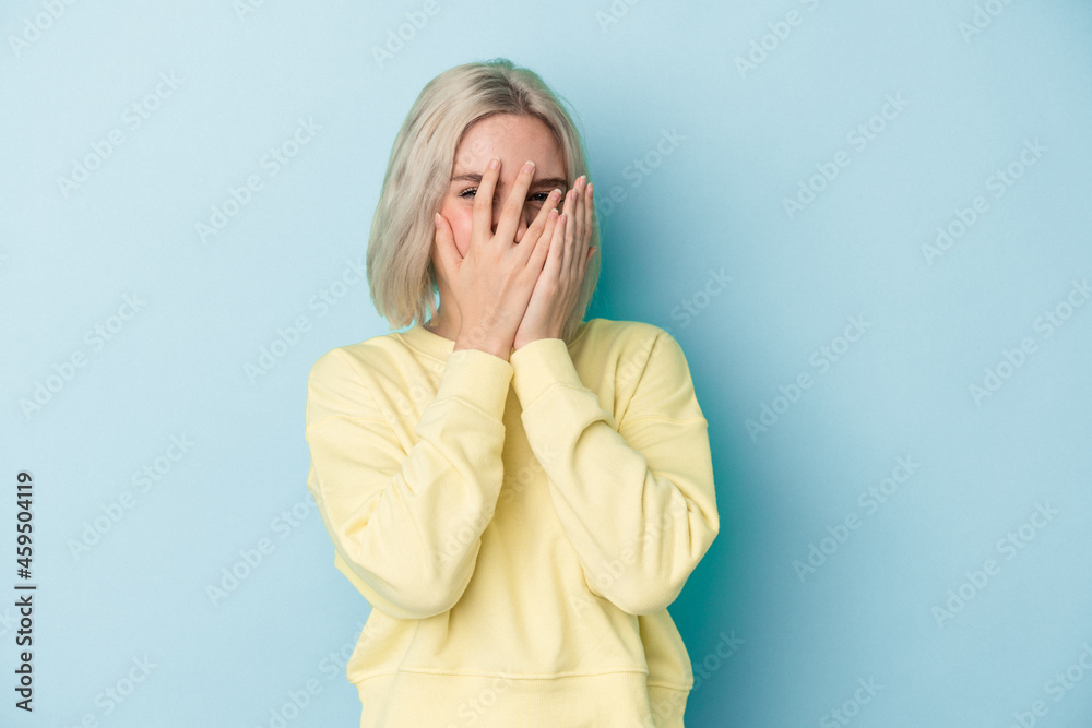 Young caucasian woman isolated on blue background blink at the camera through fingers, embarrassed covering face. Young caucasian woman isolated on blue background blink at the camera through fingers, embarrassed covering face.