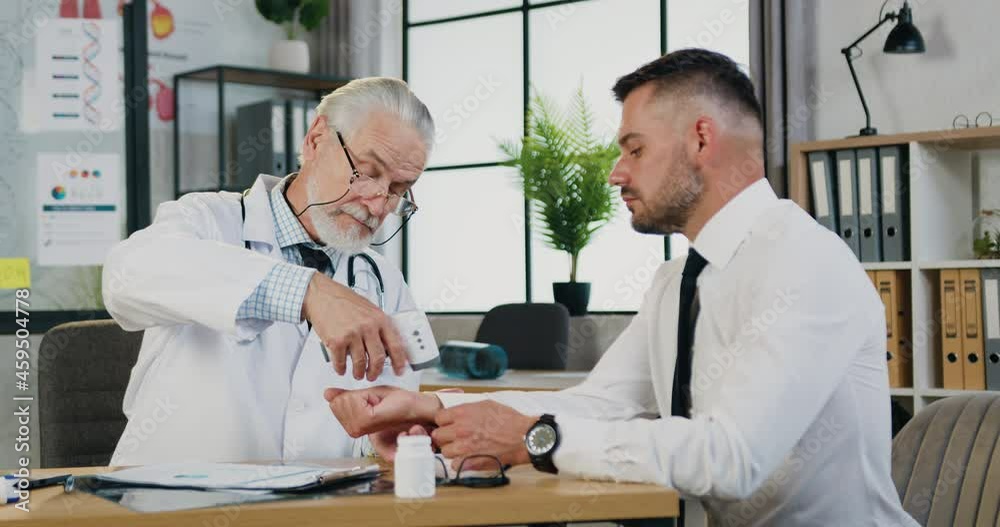 Mature doctor measuring temperature of sick patient with infrared thermometer in private hospital or clinic during COVID-19 pandemic Mature doctor measuring temperature of sick patient with infrared thermometer in private hospital or clinic during COVID-19 pandemic
