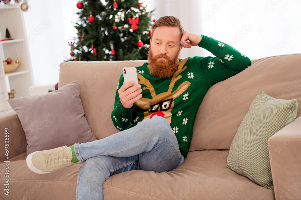 Photo of young man sit couch happy positive smile look read browse cellphone christmas eve time indoors Photo of young man sit couch happy positive smile look read browse cellphone christmas eve time indoors