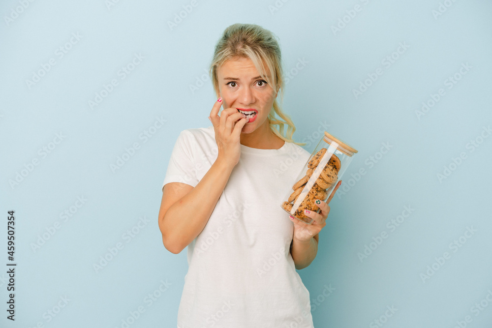 Young Russian woman holding a cookies isolated on blue background biting fingernails, nervous and very anxious. Young Russian woman holding a cookies isolated on blue background biting fingernails, nervous and very anxious.