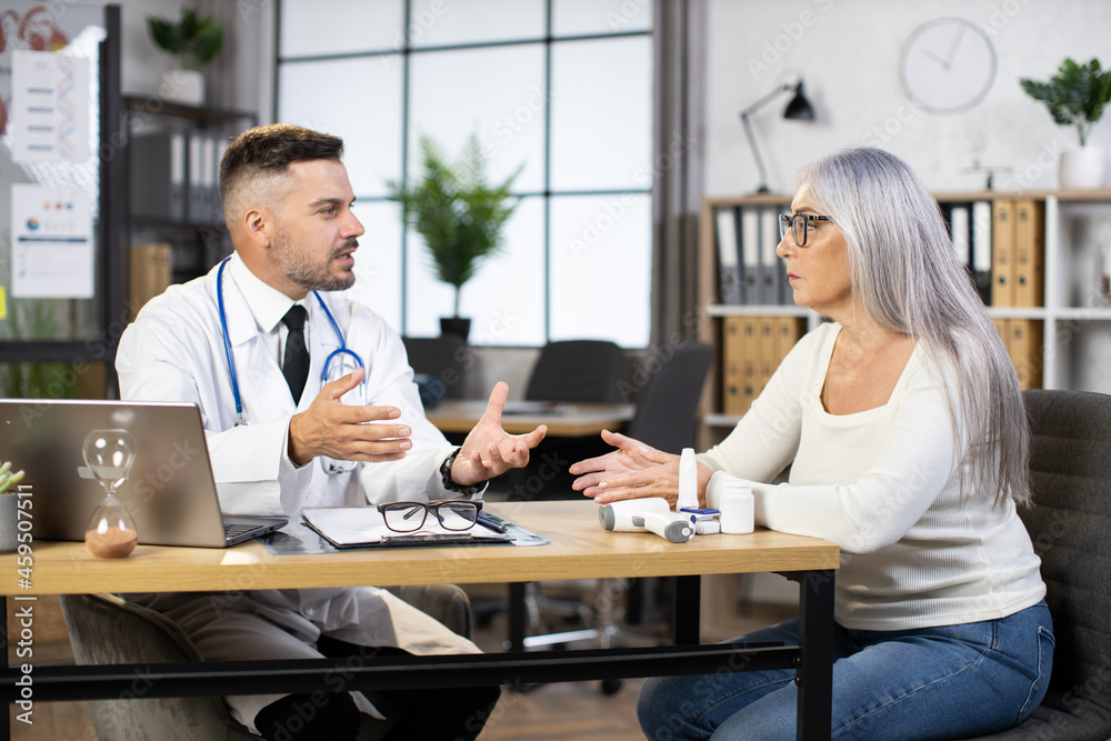 Caucasian mature woman in casual wear having regular appointment at gp physician. Male practitioner in white lab coat sitting at desk with laptop and talking with female patient. Caucasian mature woman in casual wear having regular appointment at gp physician. Male practitioner in white lab coat sitting at desk with laptop and talking with female patient.