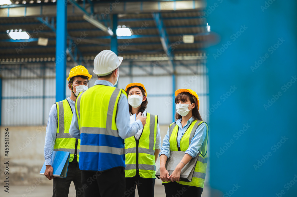 The factory employee consists of engineers, foreman, technicians, and related department staff. Wear a mask, hard hat, and vest. meeting before starting work inside the warehouse. Teamwork concept. The factory employee consists of engineers, foreman, technicians, and related department staff. Wear a mask, hard hat, and vest. meeting before starting work inside the warehouse. Teamwork concept.