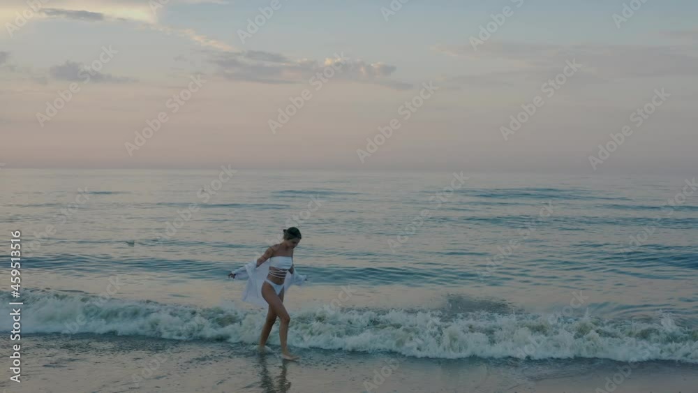 A young beautiful girl in a white swimsuit walks along the seashore at morning dawn. Thunderclouds over the sea. White sand. The sun's rays are visible through the clouds. A young beautiful girl in a white swimsuit walks along the seashore at morning dawn. Thunderclouds over the sea. White sand. The sun's rays are visible through the clouds.
