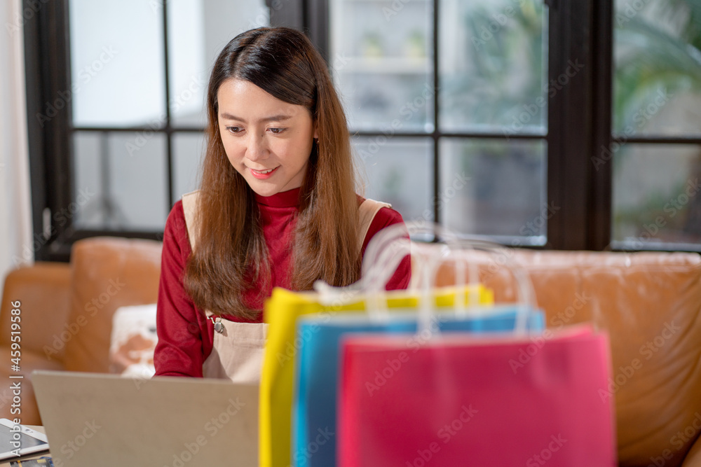 Young Asian woman use laptop for online sale business with colorful bags as foreground with concept of small business for young generation and new modern lifestyle.