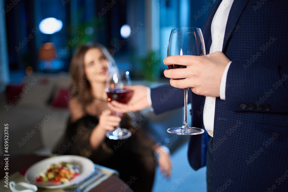Beautiful young couple in elegant wear spending evening time at home for romantic dinner. Man giving glass of red wine to charming woman. Close up. Beautiful young couple in elegant wear spending evening time at home for romantic dinner. Man giving glass of red wine to charming woman. Close up.