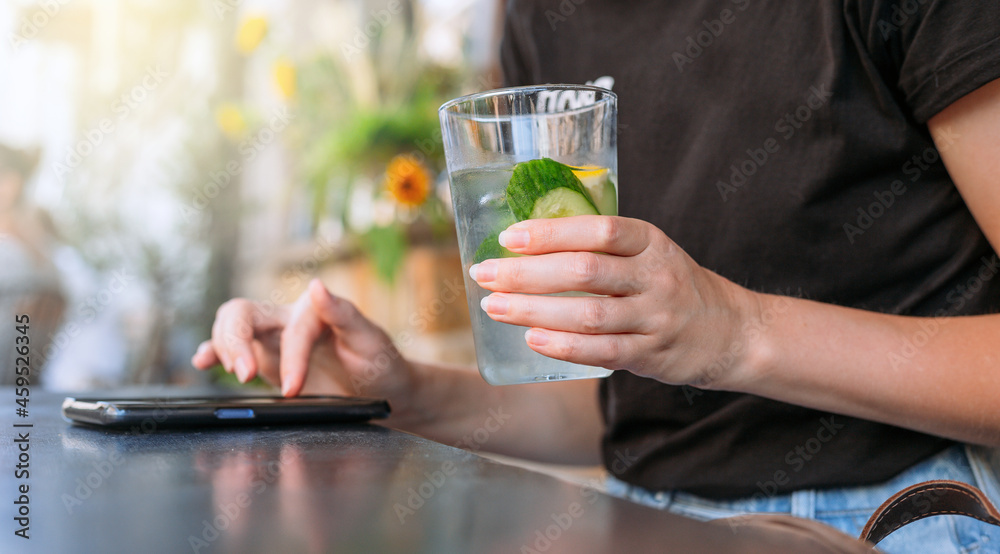 Woman at cafe drinking water and using mobile phone. Hand holding lemonade on cafe background. Glass of Cold drinks. Street front view, girl waiting for her order Woman at cafe drinking water and using mobile phone. Hand holding lemonade on cafe background. Glass of Cold drinks. Street front view, girl waiting for her order