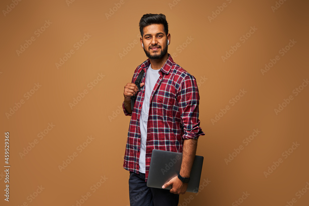 © Тарас Нагирняк - Smiling indian man in casual close with laptop and backpack on pastel background © Тарас Нагирняк - Smiling indian man in casual close with laptop and backpack on pastel background