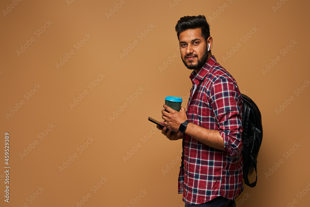 Handsome hindu man looking to camera with coffe to go and backpack on pastel background copy space Handsome hindu man looking to camera with coffe to go and backpack on pastel background copy space