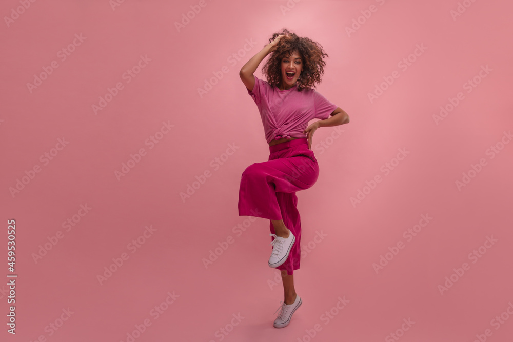 Optimistic young swarthy lady bounces on one leg with copy of space. Vibrant full-length studio photo of energetic woman wearing T-shirt and pants. Beauty, people emotions and summer vacation concept.