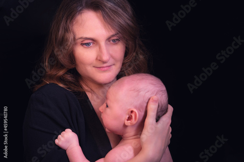 Happy woman mother holds a newborn baby boy in her arms, black studio background. Mom smiles with a child in her hands
