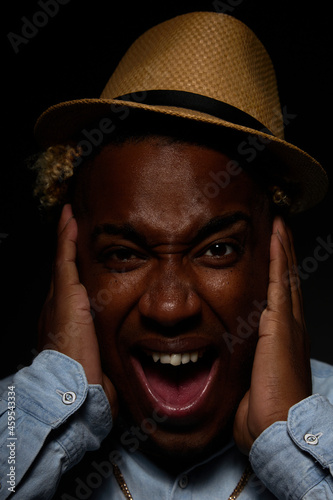 African-American man in a straw hat and a blue shirt, crazy and scared, holds his head in his hands with his mouth open from shock on a black background