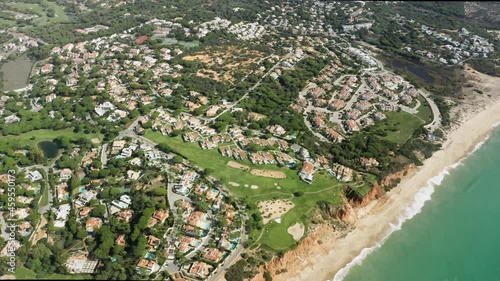 Drone shot of Faro city located in green area at Atlantic ocean, Portugal, Europe. Aerial view of residential houses surrounded parks in summer time. Sea waves on sandy beach, 4k footage