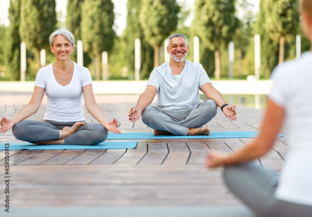 Senior couple staying in lotus pose in front of female yoga trainer during yoga class in city park Senior couple staying in lotus pose in front of female yoga trainer during yoga class in city park