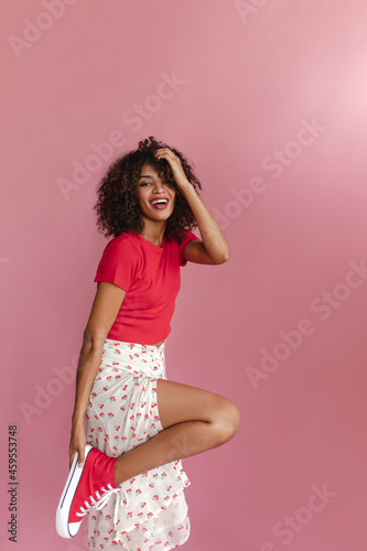 Young African woman stands half sideways and bends leg to knee on pink background. Softly touching curly and fluffy dark blond hair with thin hand. Looks beautiful in red T-shirt and skirt with slit. © Look!