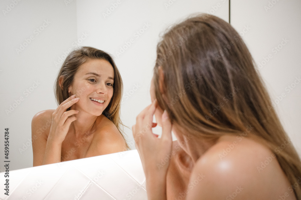 young white woman in the bathroom mirror looking at skin looking for spots signs of age and lines happily and confident in the bathroom after shower self care skin care anti aging young adult young white woman in the bathroom mirror looking at skin looking for spots signs of age and lines happily and confident in the bathroom after shower self care skin care anti aging young adult