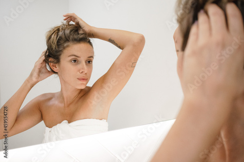 young white woman bathroom mirror messy bun updo fixing hair after shower self care skin care routine getting ready at home happy confident feeling beautiful and attractive in love with herself