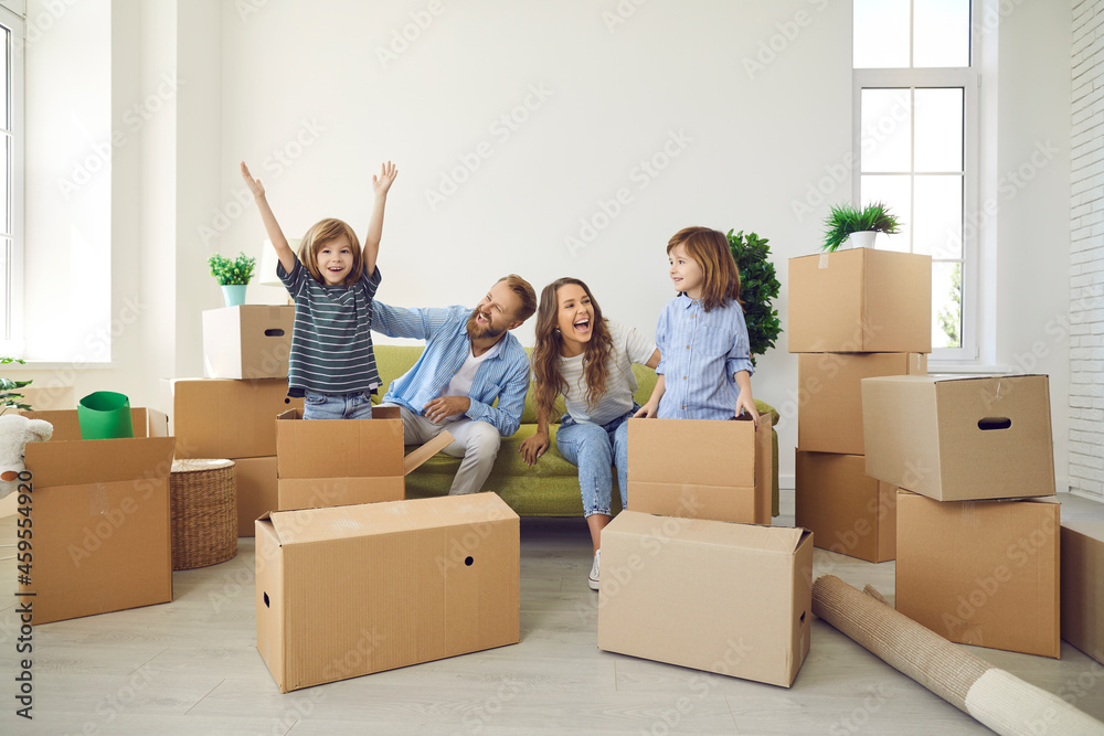 Happy family having fun in new home. Cheerful young mommy and daddy sitting on sofa and laughing while excited little children are playing with cardboard boxes in spacious living room in new apartment Happy family having fun in new home. Cheerful young mommy and daddy sitting on sofa and laughing while excited little children are playing with cardboard boxes in spacious living room in new apartment