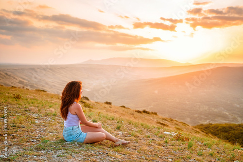 A young woman is sitting on the mountainside at sunset and enjoying the view of the mountains. The concept of an active lifestyle and peace.