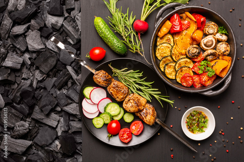 Plate with kebab and fresh vegetables, frying pan with grilled vegetables on a black wooden charcoal tabletop. Top view.