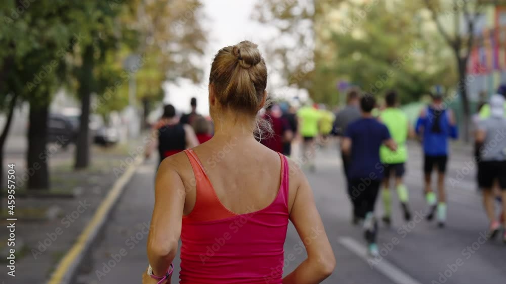 Slow motion blonde woman with hair bun wearing top and earphones running on empty city road, blurred crowd running ahead of her. Following shot of athletes taking part in sports event Slow motion blonde woman with hair bun wearing top and earphones running on empty city road, blurred crowd running ahead of her. Following shot of athletes taking part in sports event