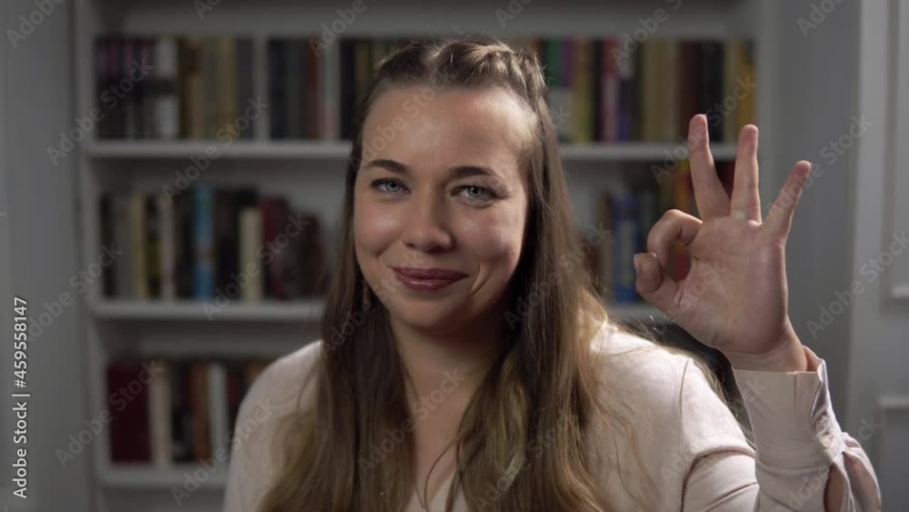 Portrait of a smiling caucasian woman showing Ok sign sitting in a room on bookshelf background. Young lady with long fair hair and blue eyes looks at camera and gestures. Concept of person's emotions