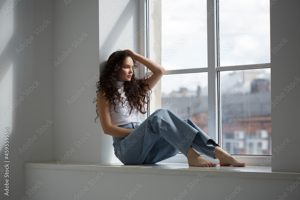 A cute slender girl with long curly hair sits at the window in a bright room on a sunny day.