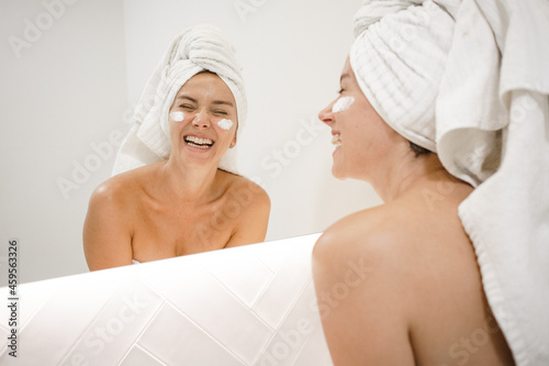 young white woman in the bathroom mirror face cream laughing anti acne anti aging spots signs of age confident in the bathroom after shower self care skin care young adult cosmetics plump toned skin