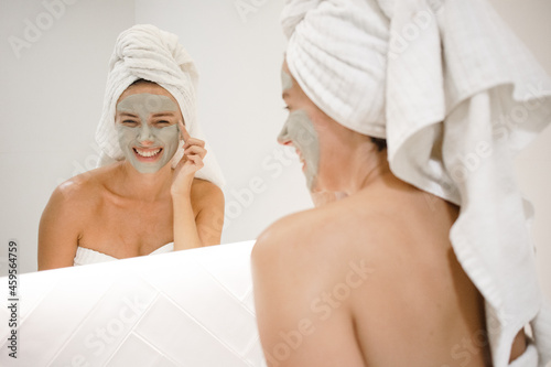 young white woman applying green clay mask bathroom mirror anti acne anti aging spots signs of age oily skin treatment after shower self care skin care young adult cosmetics plump toned spotless skin