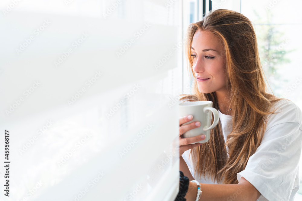 Beautiful young woman with mug looking through window at home Beautiful young woman with mug looking through window at home