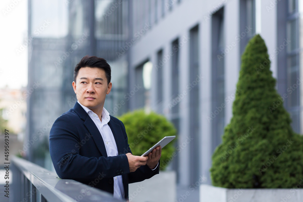 Serious asian boss reads news from tablet near modern office male businessman in business suit Serious asian boss reads news from tablet near modern office male businessman in business suit