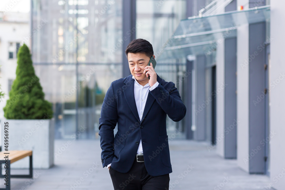 Successful Asian businessman businessman talking on a mobile phone while walking near the office of a large modern, serious and pensive employee Successful Asian businessman businessman talking on a mobile phone while walking near the office of a large modern, serious and pensive employee