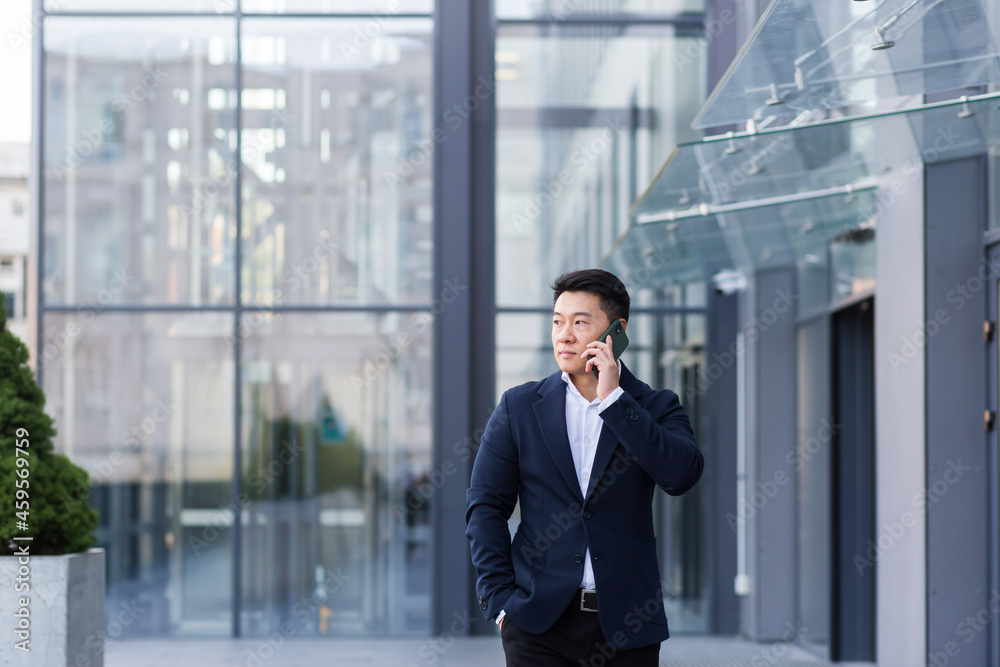 Successful Asian businessman businessman talking on a mobile phone while walking near the office of a large modern, serious and pensive employee Successful Asian businessman businessman talking on a mobile phone while walking near the office of a large modern, serious and pensive employee