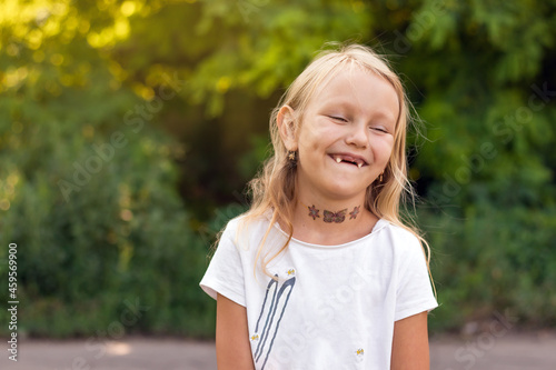 funny little girl, blonde, 6 years old, laughs cheerfully, opening her mouth without milk upper teeth, child's portrait of a toothless girl on the background of the park