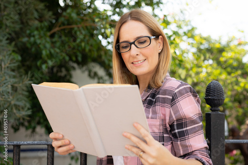 Beautiful smiling woman in casual shirt and glasses reads white book against summer green park.