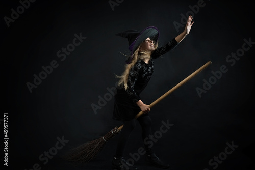 A little girl on a dark background in a witch costume and a wide-brimmed hat flies on a broom on Halloween.