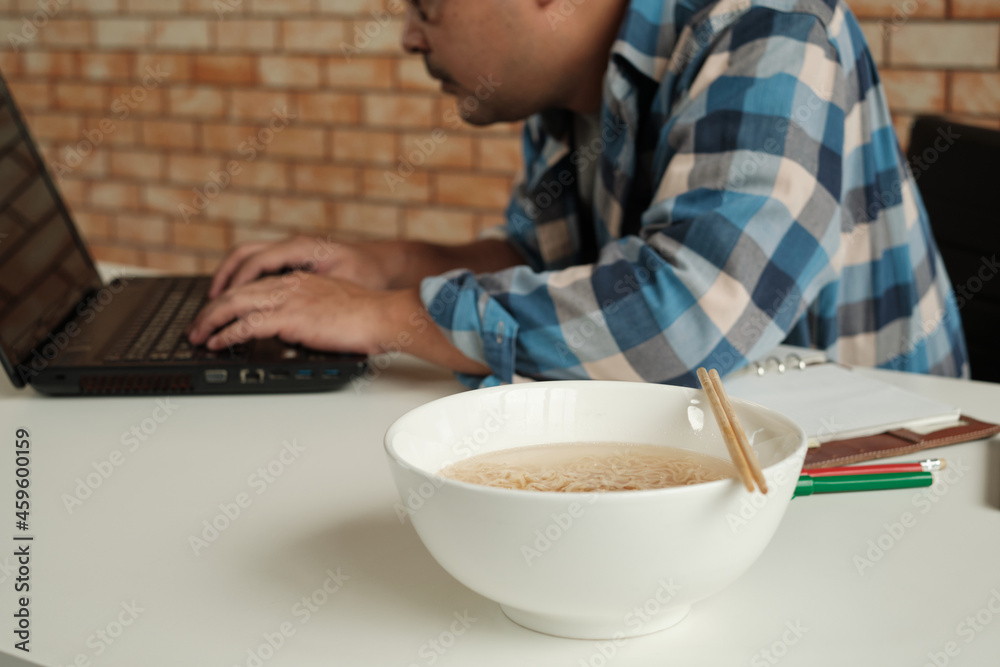 Thai male worker busy working with laptop, use chopsticks to hastily eat instant noodles during office lunch's break, because quick, tasty, and cheap. Over time Asian fast food, unhealthy lifestyle. Thai male worker busy working with laptop, use chopsticks to hastily eat instant noodles during office lunch's break, because quick, tasty, and cheap. Over time Asian fast food, unhealthy lifestyle.