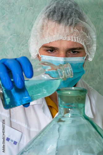 a medical laboratory assistant in a white coat gloves a hygienic mask and a cap pours liquid from one vessel to another of a larger volume